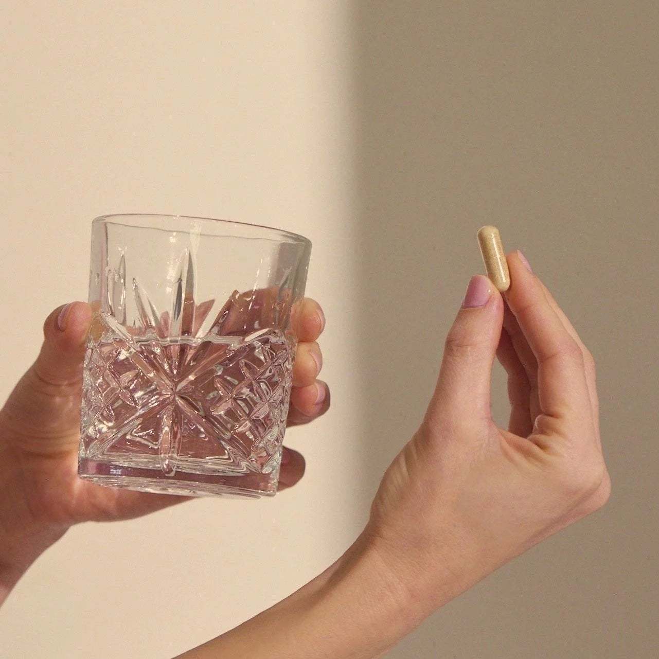 Hand holding a crystal glass with ice and a gold pill against a beige background