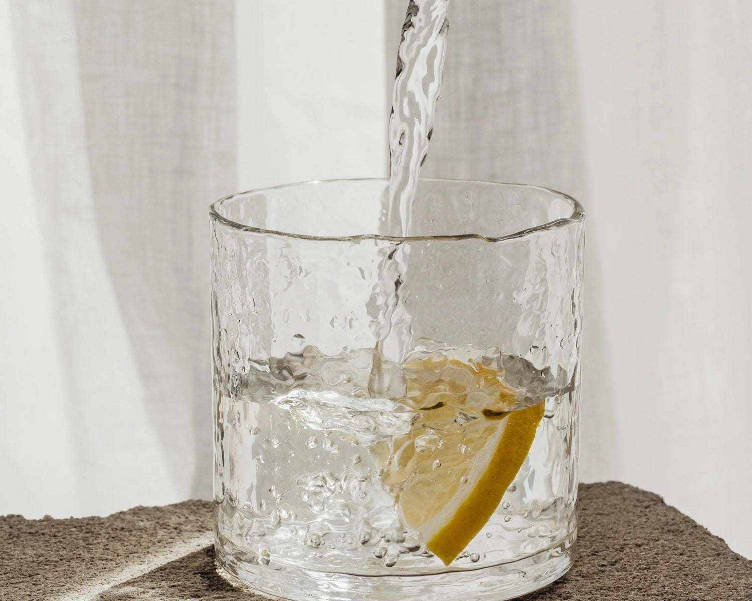 Clear glass with a lemon wedge being filled with water against a white curtain background