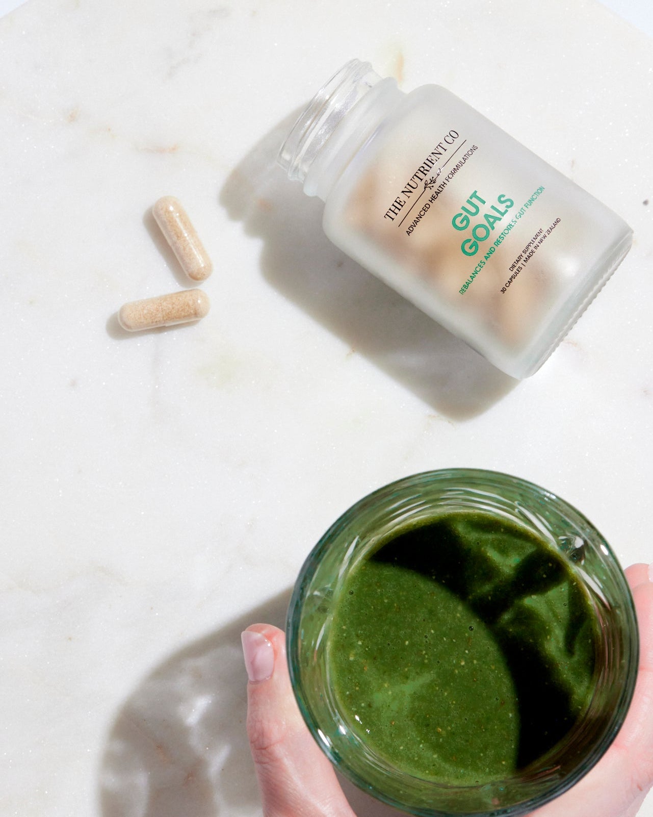 Hand holding a green smoothie with a container labeled 'Gut Goals' and capsules on a light background